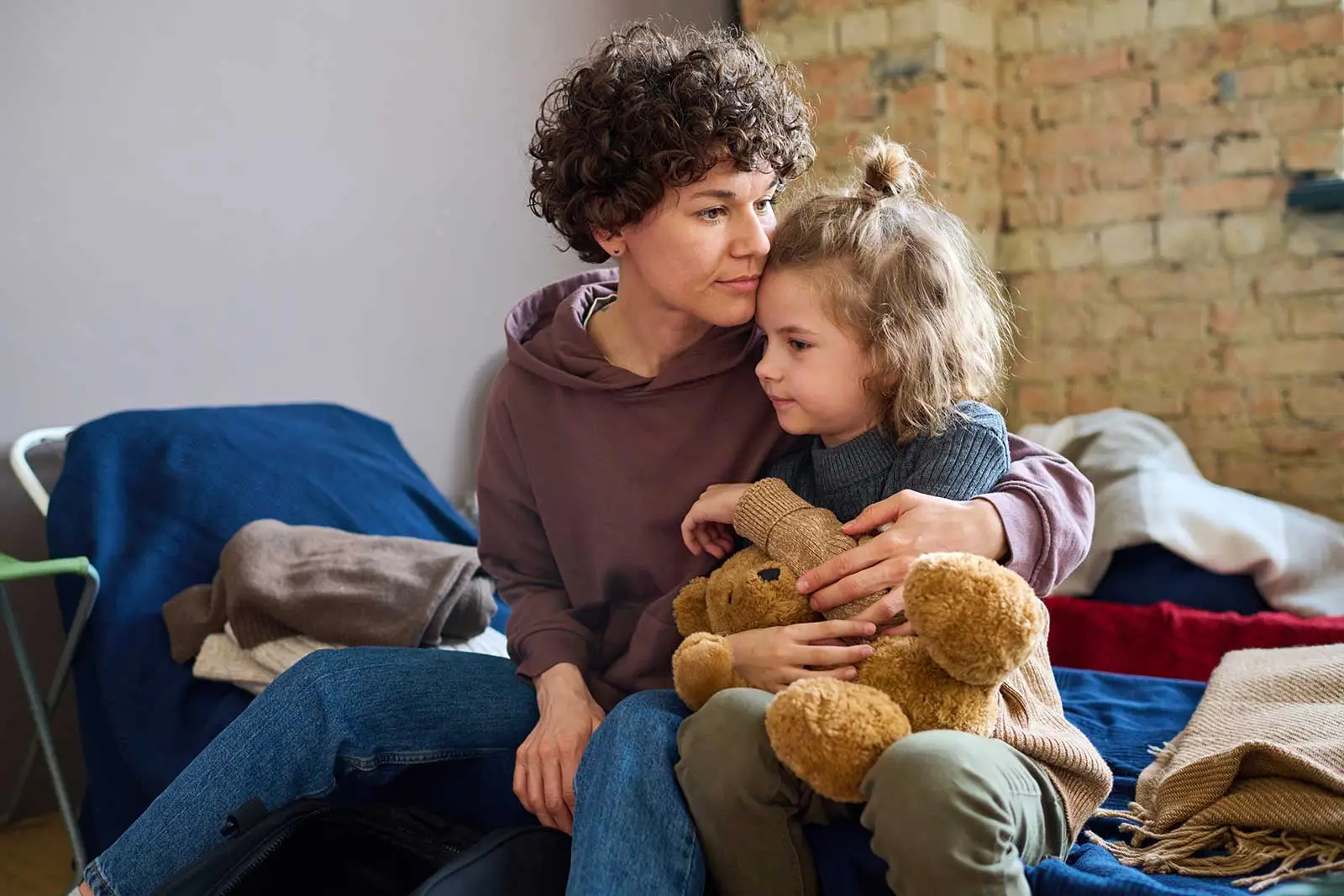 A mother sits on a makeshift camping bed looking sad cuddling her young daughter in a homeless shelter