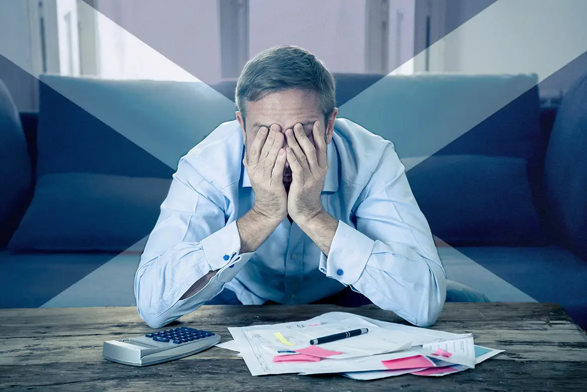 A man stressing over bills with his heads in his hands with a saltire faded in the background
