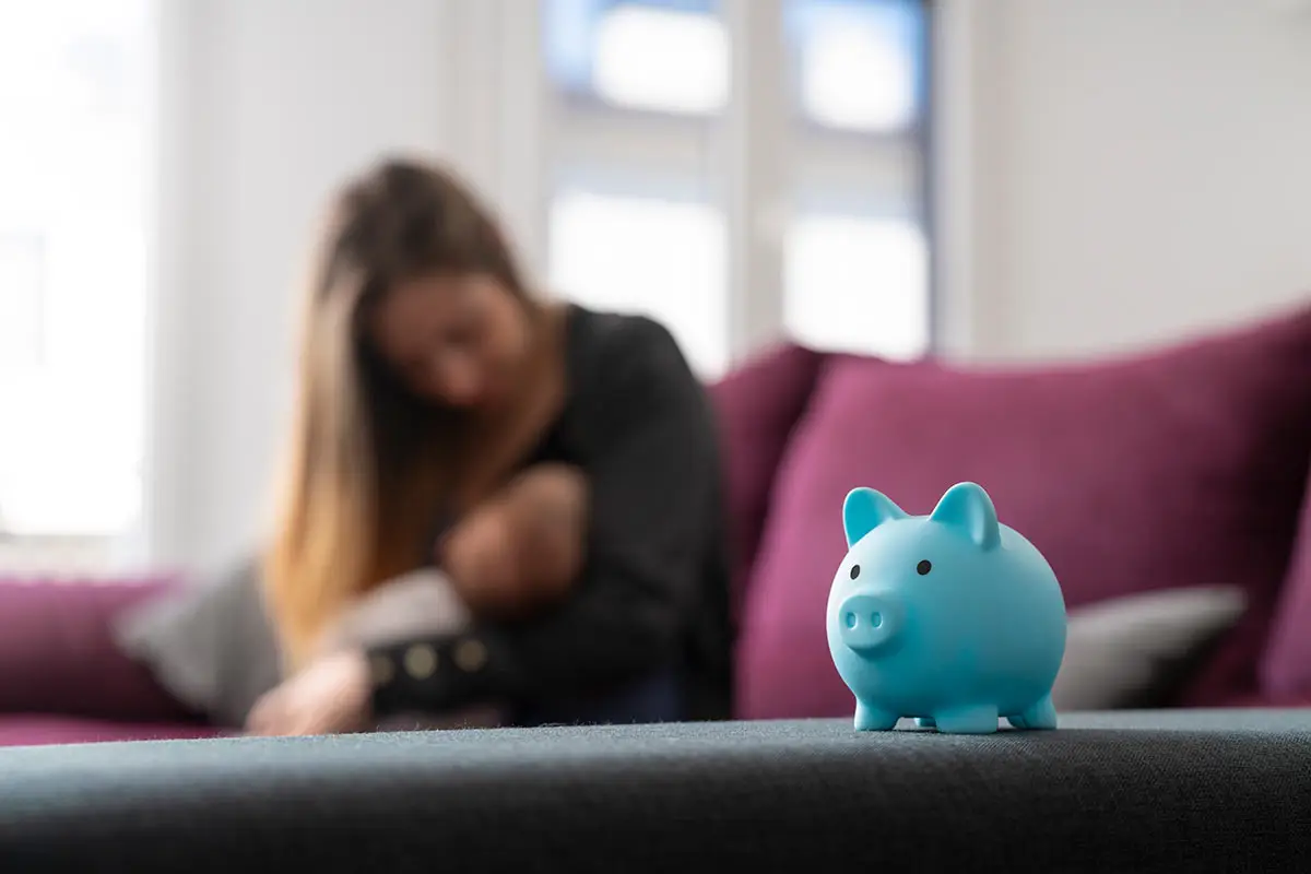Foreground image of a piggy bank with a single mother in the background with her baby